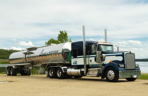 Liquids in Motion blue, white, and silver bulk liquid hauling truck on gravel road in front of a lake. 